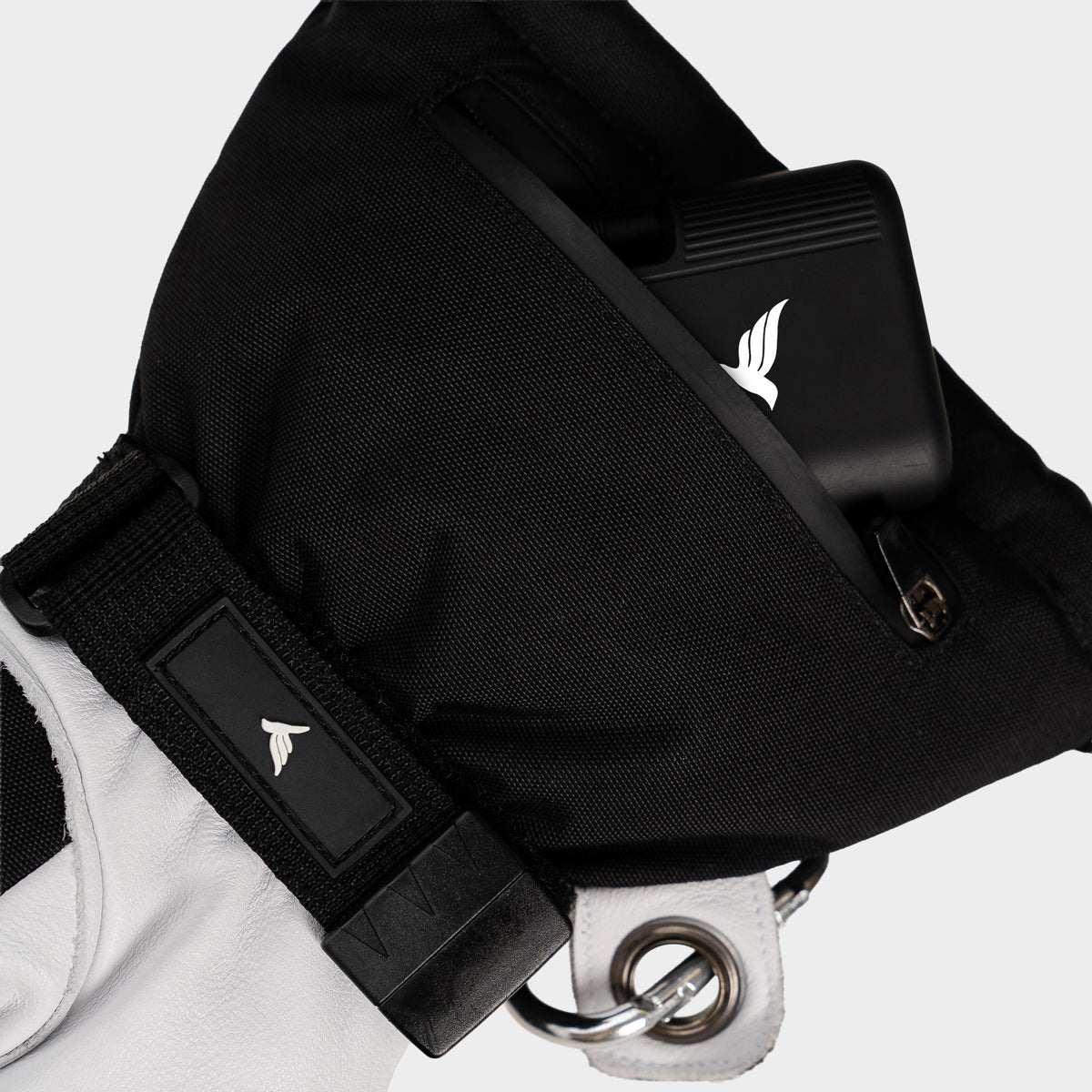Close-up of a black tactical shoulder bag with a white wing logo, shown worn over a white sleeve. The bag features a black strap with a matching logo patch and a silver metal grommet.