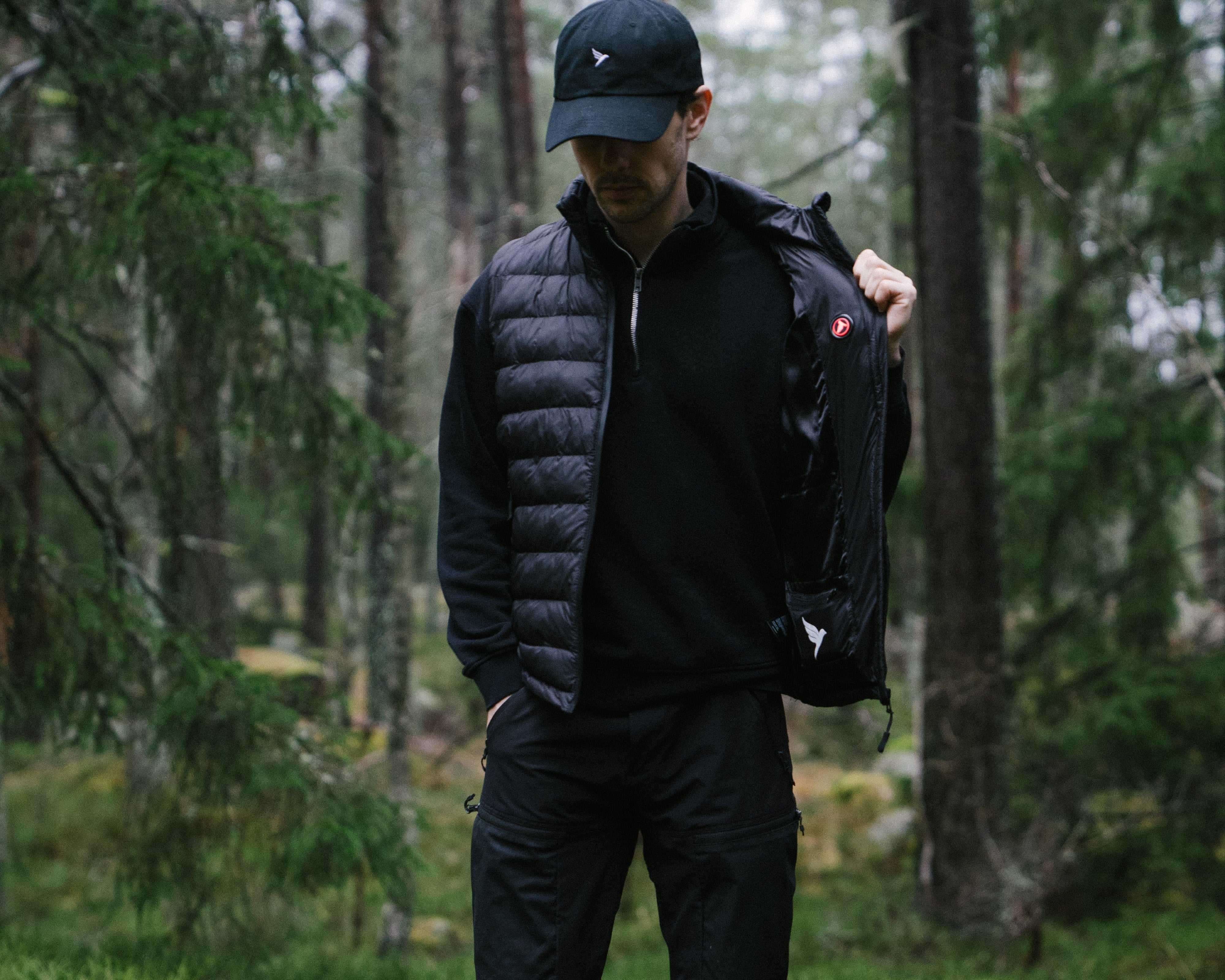 Person wearing a black heated vest and cap in a forest setting