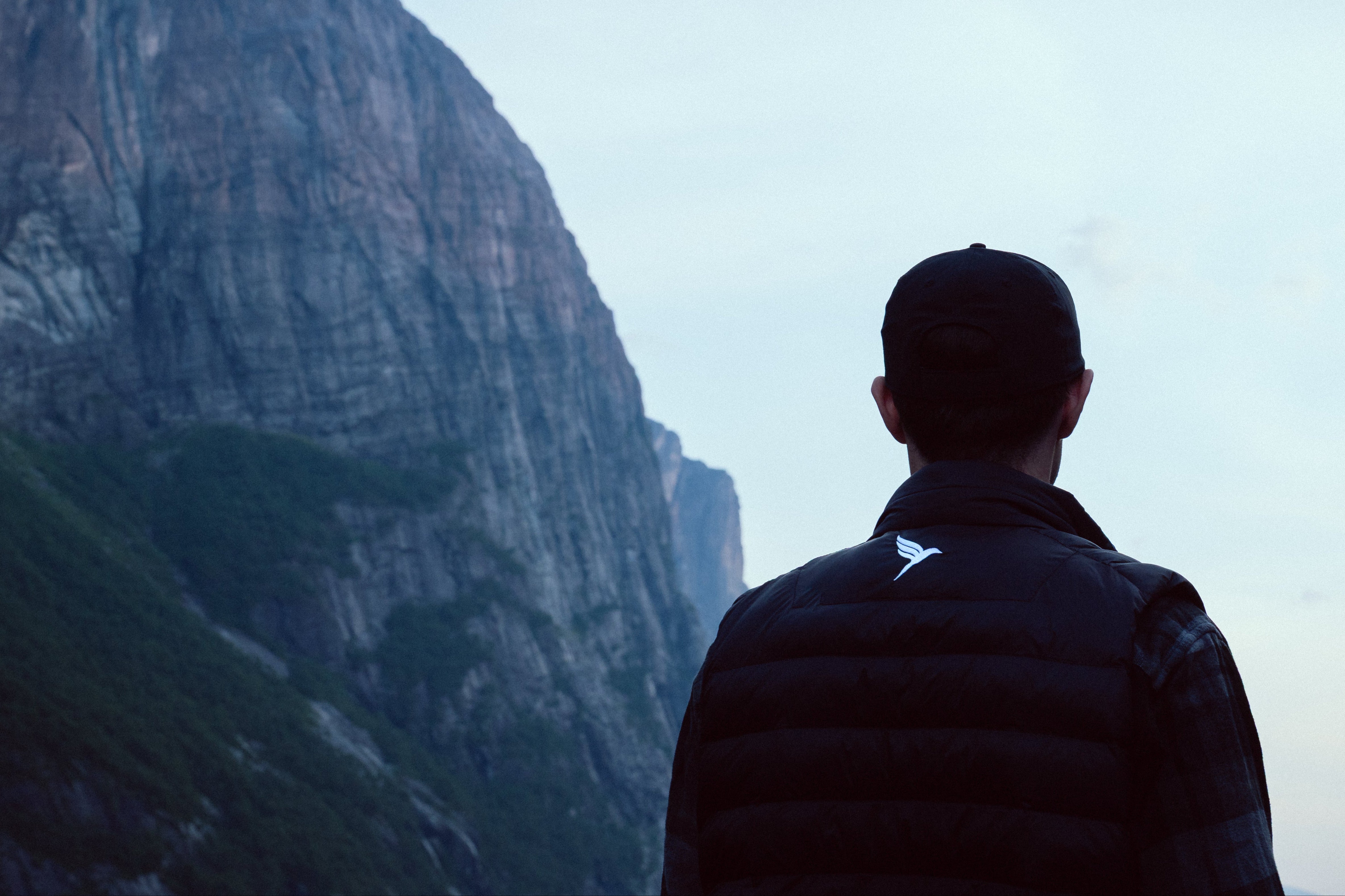 Person wearing a black jacket with a logo, standing in front of a mountainous landscape.