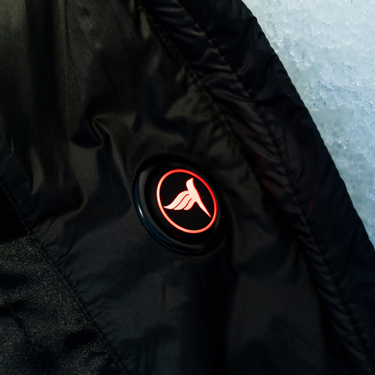 Close-up of a black puffer jacket fabric with a circular black patch featuring a red illuminated logo, next to a light icy blue surface.